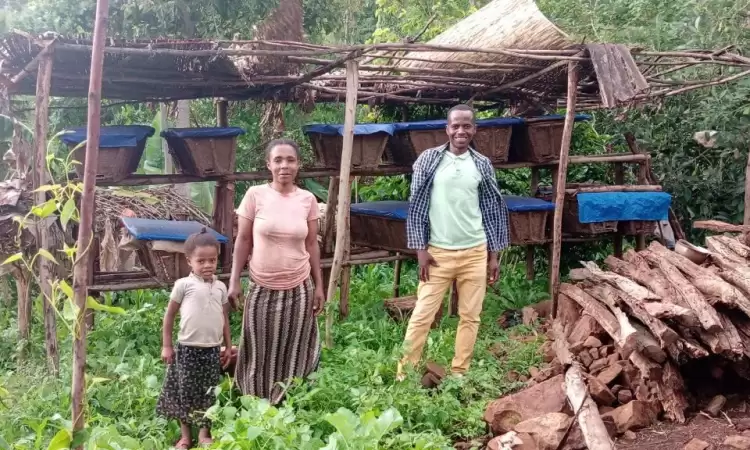Ethiopian man standing in his field next to his homemade beehives, with his wife and daughter