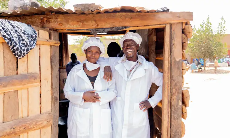 A Ugandan man and woman stand in front of their bakery, in white cooking uniforms, smiling to the camera
