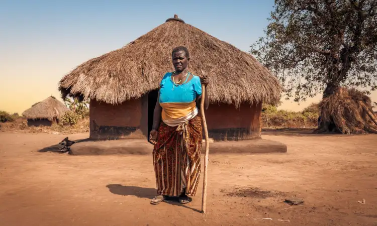 A Ugandan woman who lives with a disability stands in front of her hut, using a stick to support her