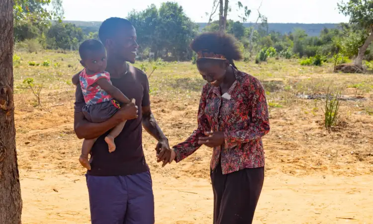 Tyson, with his wife and young child on their dried-out farm. He is holding his child, and his wife's hand, and they are laughing.in Kenya