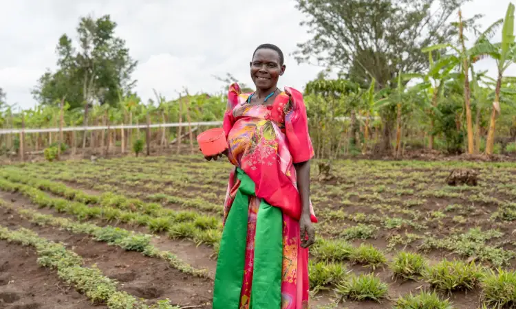 A Ugandan woman, wearing a pink and green floral dress, is standing in her field smiling, her hand muddy from planting seeds