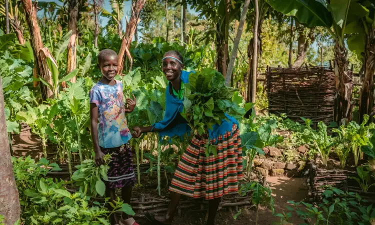 A Kenyan woman and her daughter, stood in their green and lush kitchen garden, a combination of crops and small wooden structures, smiling at the camera and holding leafy greens
