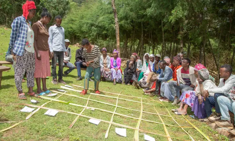 Ethiopian family participating in THM session, placing stones in the grid made of bamboo on the floor, community members are sat around and watching