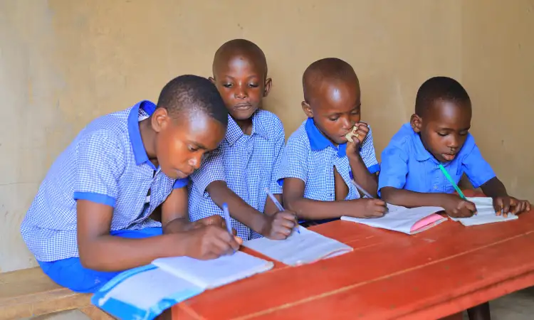 Four children dressed in light blue uniforms, sat at a table and writing in their notebooks