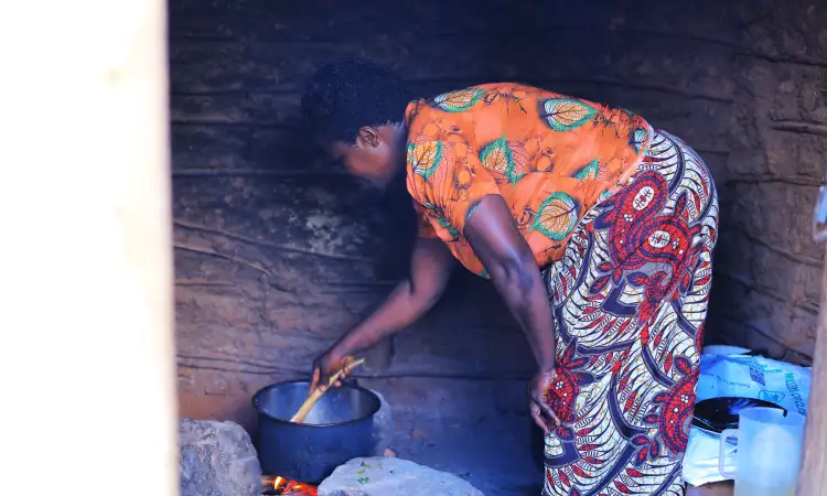 A woman, Jeanne, is dressed in colourful orange and burgundy, and is bending over her cooking pot, stirring its contents