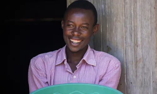 A young man smiling and holding a large green basin filled with harvested grains at the entrance of a wooden storage hut.