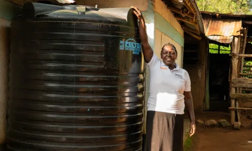 Florah with her hand on her large water tank, smiling