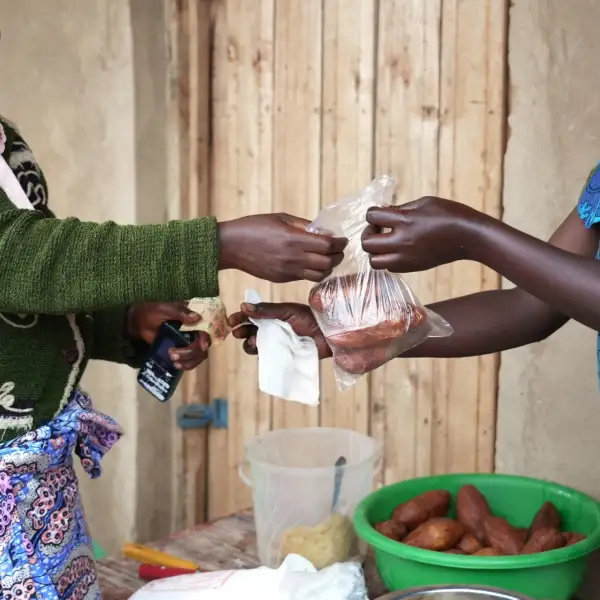 Two women exchanging money and packaged food during a small-scale food sale outside a rural home.