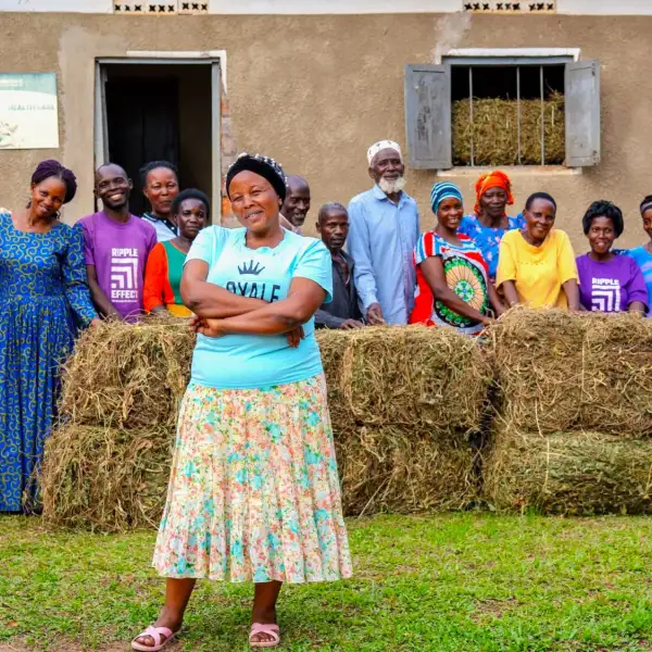 A women-led farmer group standing proudly in front of stacked hay bales, smiling for a group photo outside a community building.